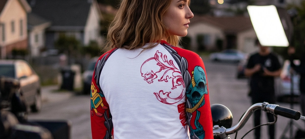 Woman wearing a white raglan T-shirt with colorful artwork beside a motorcycle at sunset