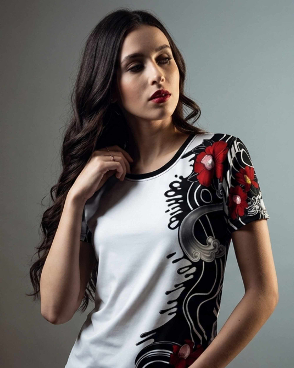 A woman modeling a white short-sleeve t-shirt with a black crew neck and a black, red, and gray graphic print on the front and sleeve, set against a plain gray studio background.