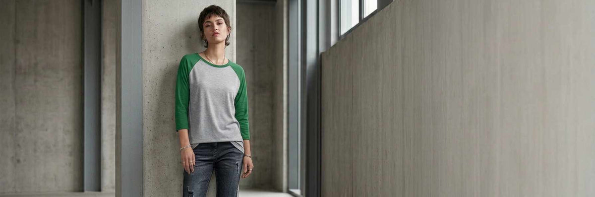 A woman wearing a three-quarter sleeve raglan t-shirt with a gray body and green sleeves, paired with dark jeans, standing in a modern concrete hallway.