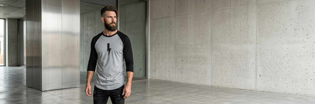 A bearded man wearing a gray and black raglan baseball t-shirt with a black graphic logo on the chest, standing in a modern concrete-walled corridor.