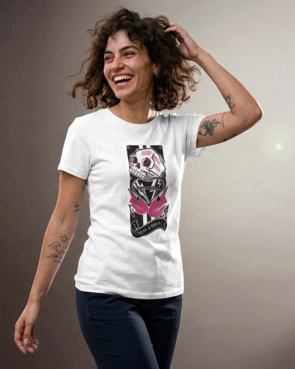 A woman with curly brown hair is smiling and laughing while wearing a white short-sleeve t-shirt with a graphic print of a skull in a black helmet with pink butterfly wings. The background is a plain gray studio wall.