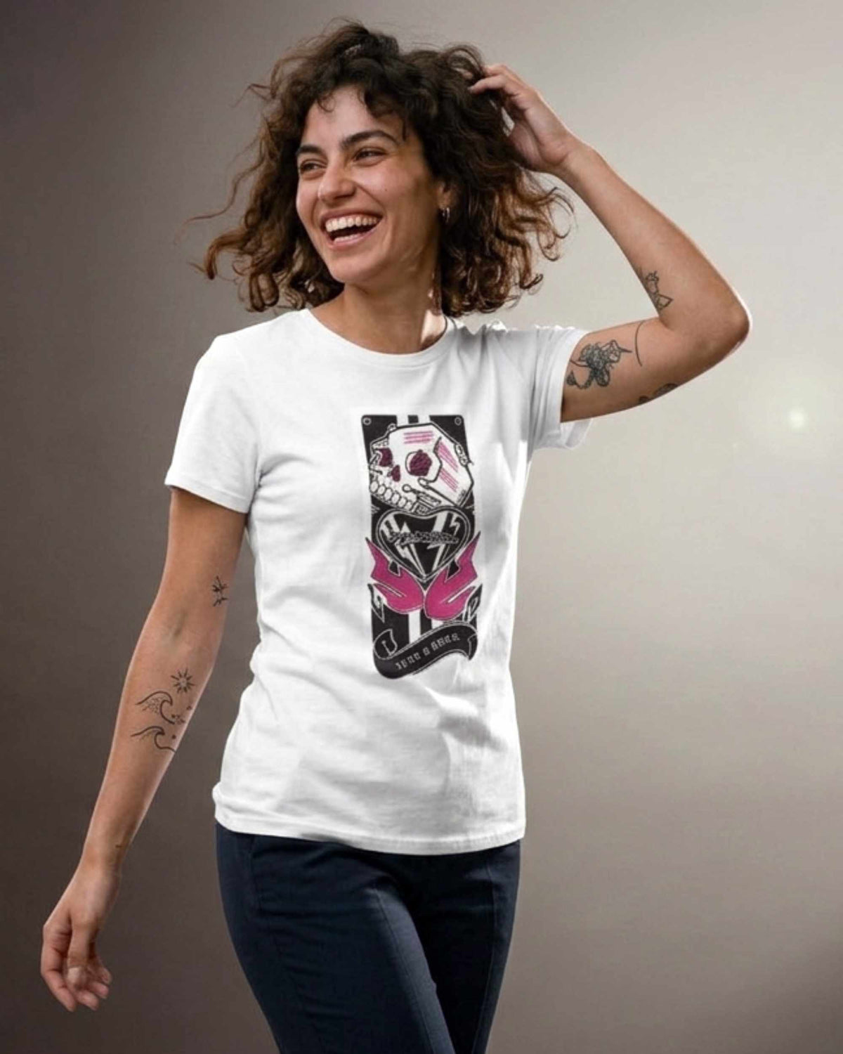 A woman with curly brown hair is smiling and laughing while wearing a white short-sleeve t-shirt with a graphic print of a skull in a black helmet with pink butterfly wings. The background is a plain gray studio wall.