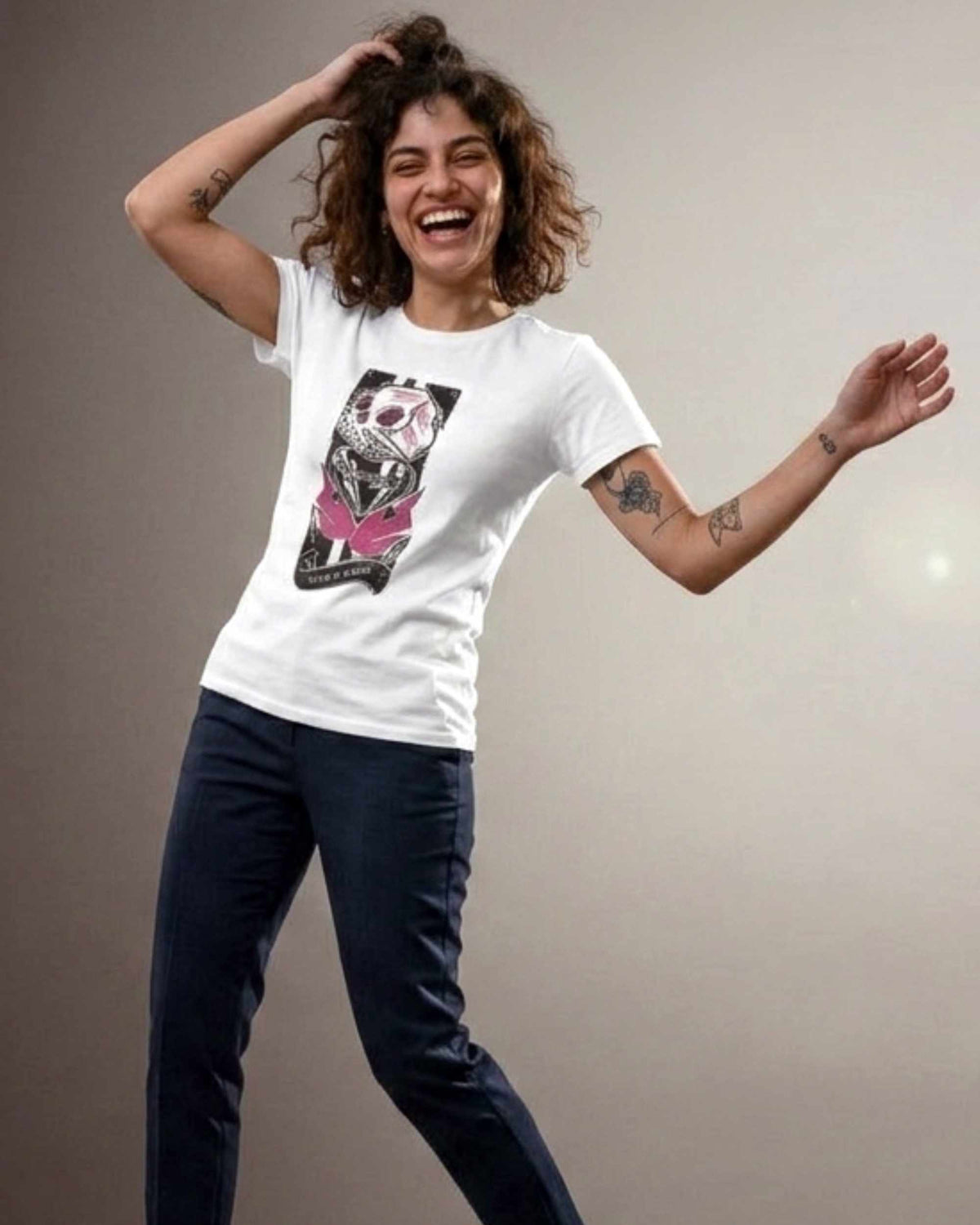 A woman wearing a white crewneck t-shirt with a black and pink graphic print of a skull, paired with dark blue jeans, posing against a neutral studio background.