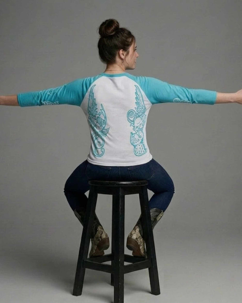 A woman seated on a black stool, photographed from the back, wearing a white and turquoise raglan baseball-style shirt with turquoise wing graphics on the back, dark blue jeans, and cowboy boots against a plain gray studio background.