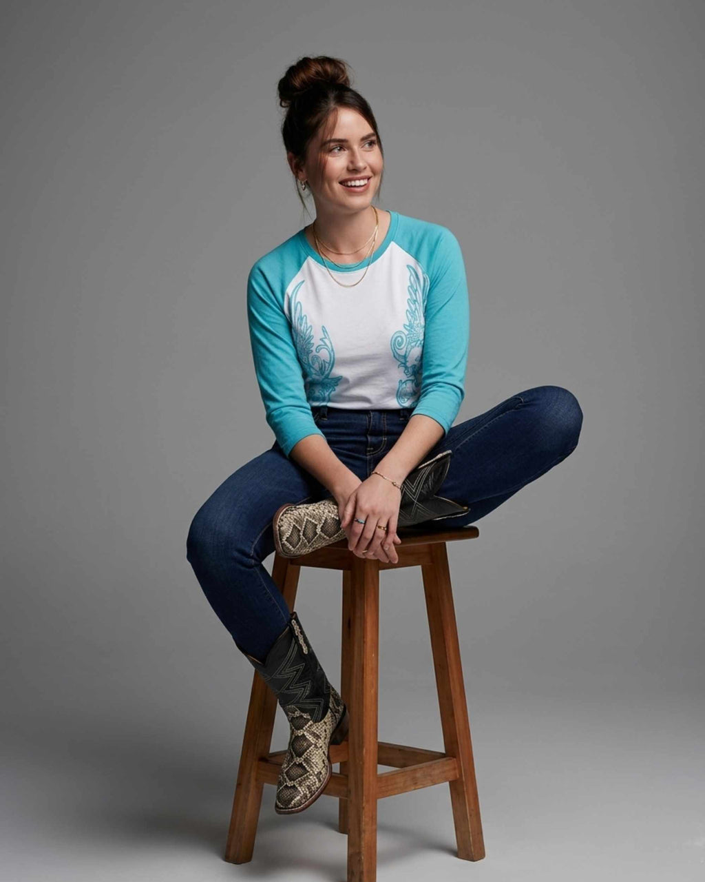 A woman sits on a wooden stool against a gray studio background, wearing a turquoise and white raglan-style long-sleeve top with blue jeans and patterned cowboy boots.