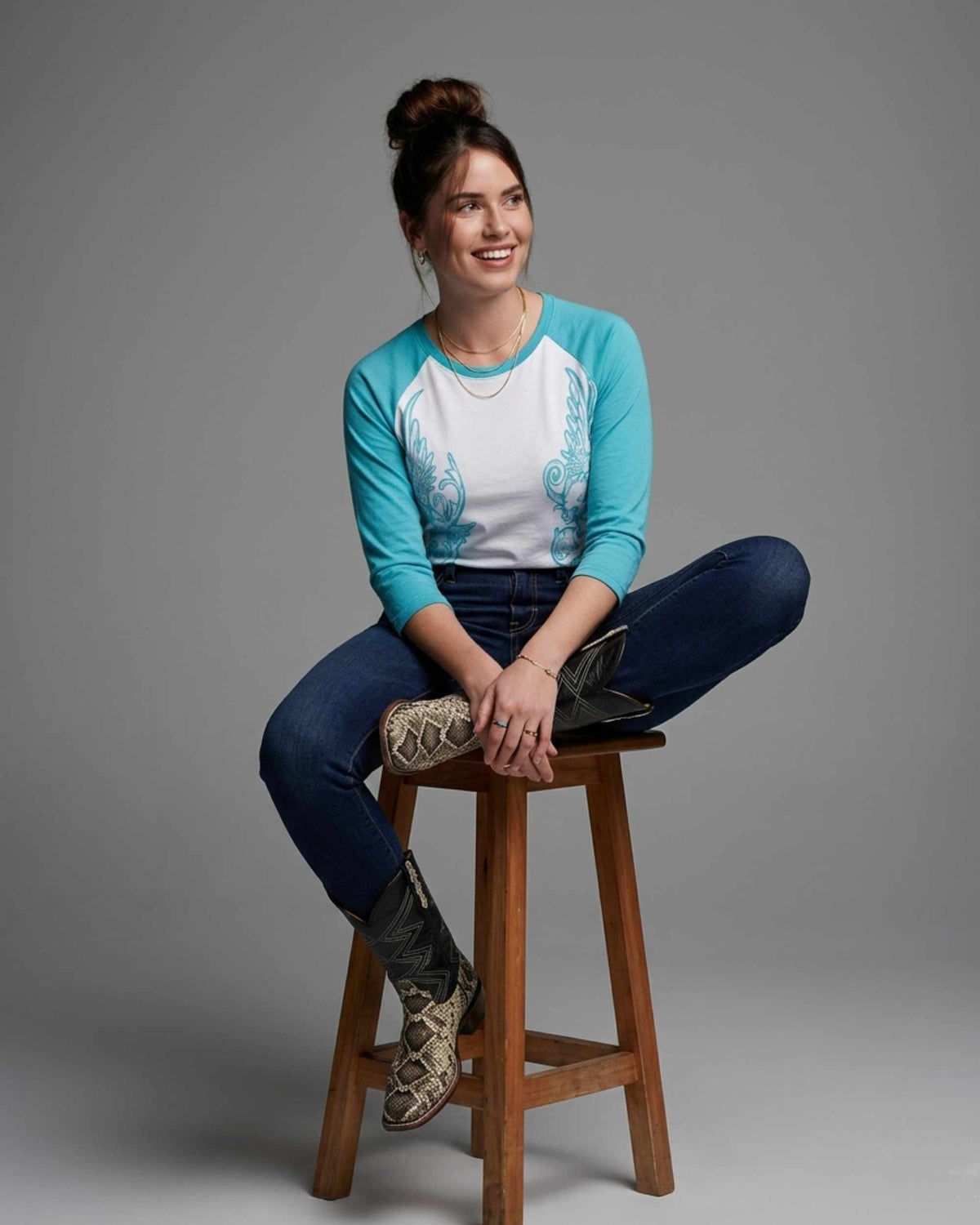 A woman sits on a wooden stool against a gray studio background, wearing a turquoise and white raglan-style long-sleeve top with blue jeans and patterned cowboy boots.