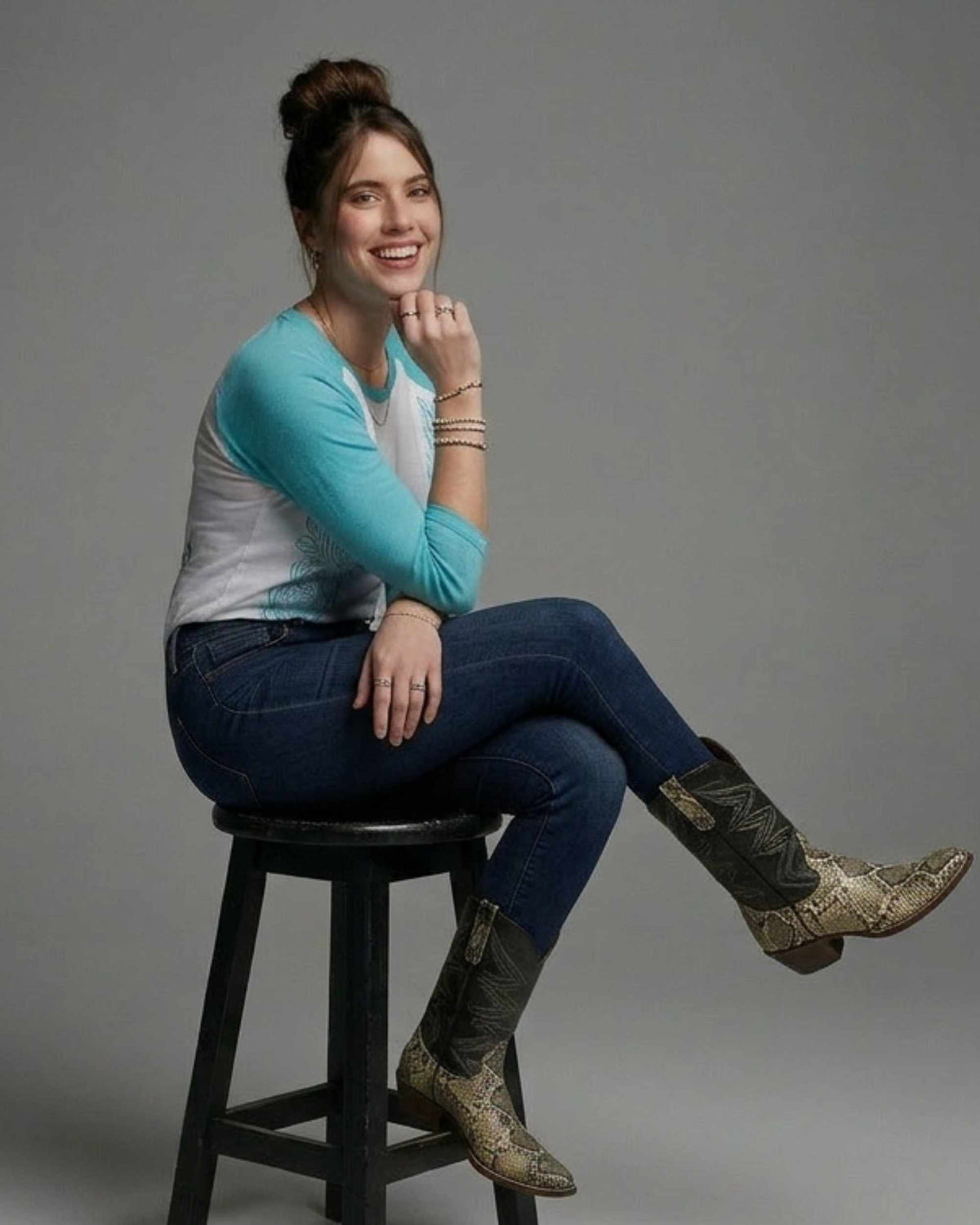 A woman sits on a black stool against a gray studio background, wearing a turquoise and white raglan long-sleeve top, dark blue jeans, and snakeskin-patterned cowboy boots.